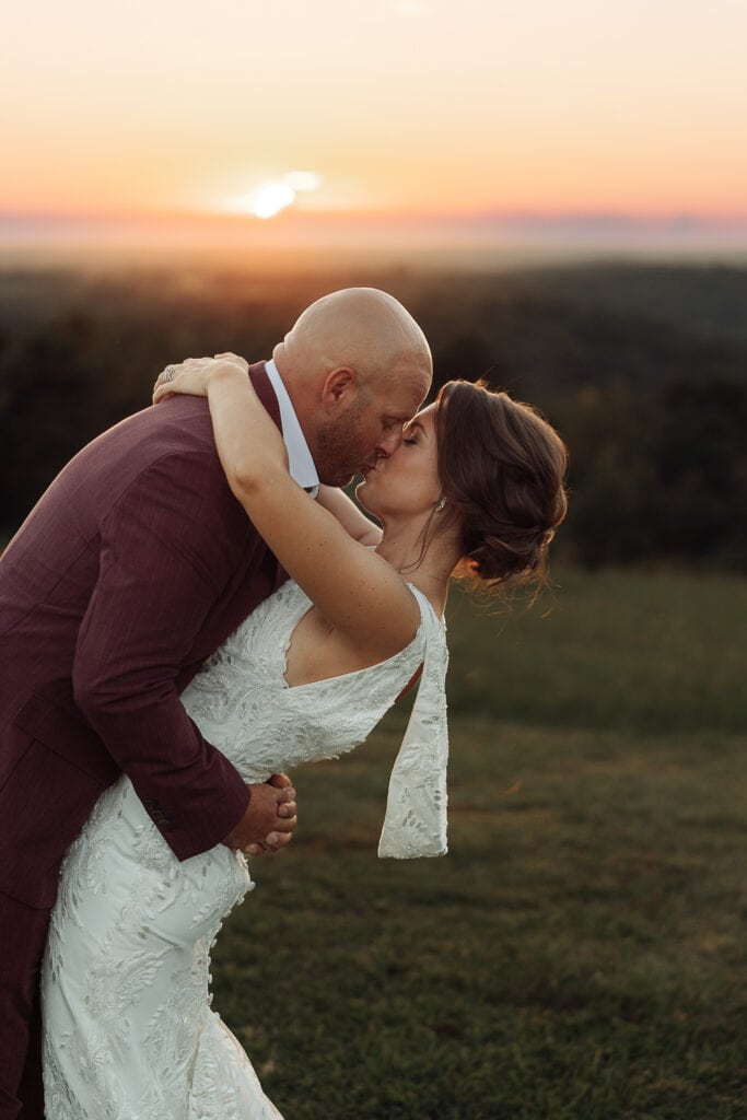 A bride and groom in a romantic embrace kiss as the sun sets behind them.