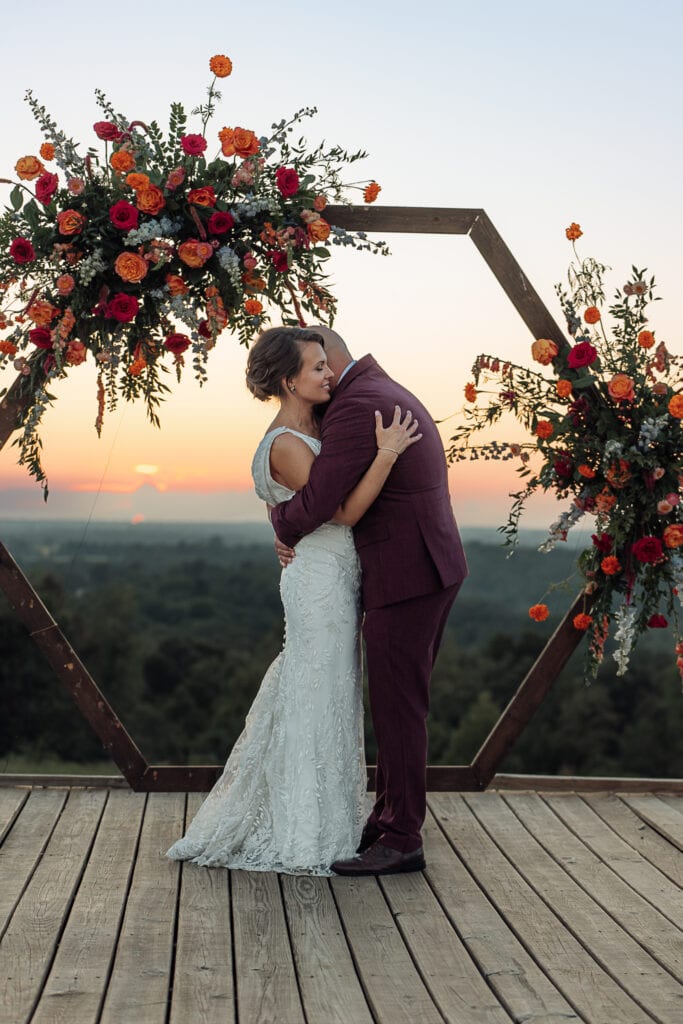 AJ hugs Lauren on a wooden platform. There is a wooden arch full of vibrant flowers behind them, and the sunset is beautiful.