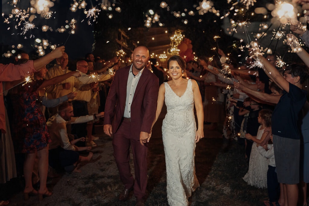 Lauren & AJ walk through a sparkler exit at their wedding. The rustic barn where they had their reception is in the background.