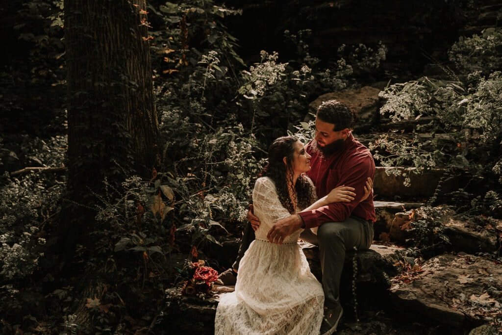 Bride and groom sit on rock steps in the forest with their arms around each other, as the sunlight shines on them through the trees.