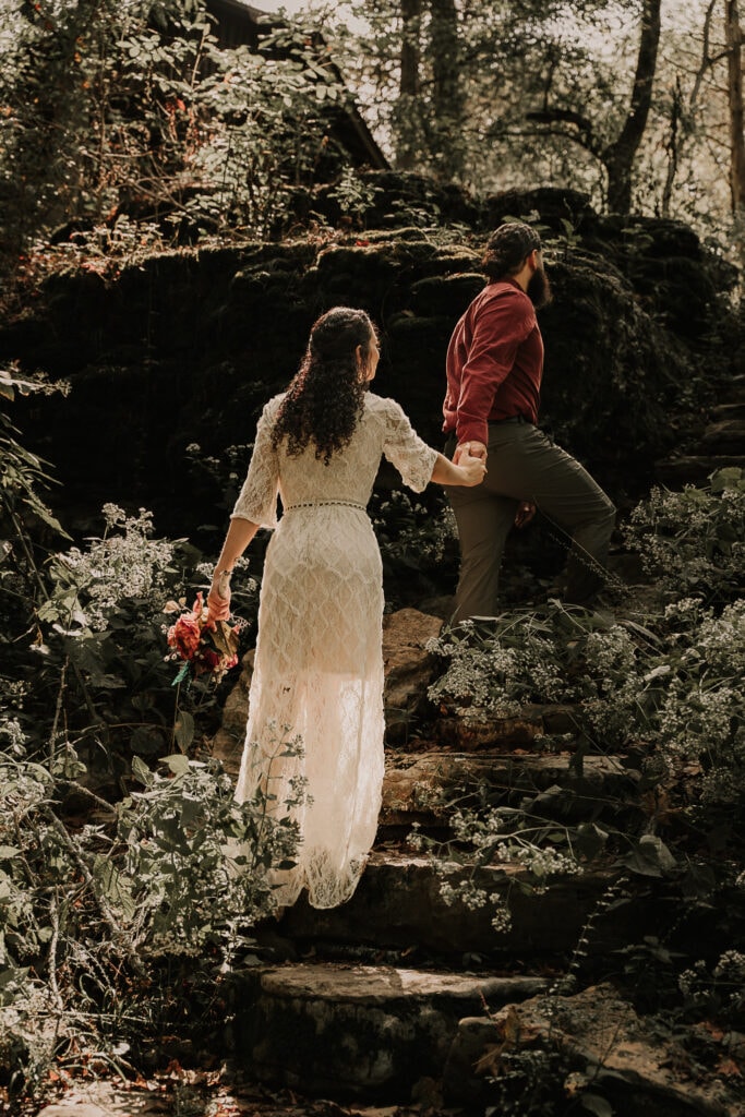 Bride and groom walk hand in hand up rock steps in the forest during their elopement. Sun shines through the trees.