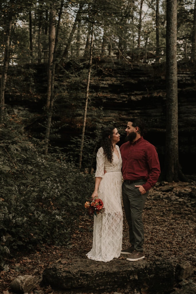 Bride and groom stand outside on a rock, facing each other. She holds her bouquet at her side, his hand is in his pocket. A cave is behind them.