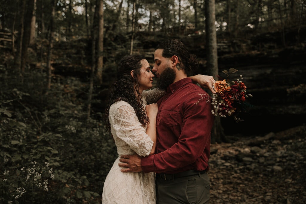 Bride and groom are in the forest, facing each other with their foreheads touching.