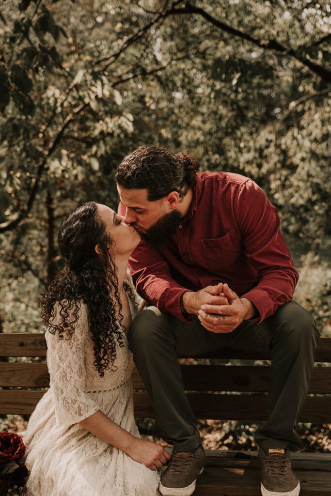 Ezekiel sits on a bench with his feet on the seat. Mady sits next to him as they kiss. They're wearing wedding attire.