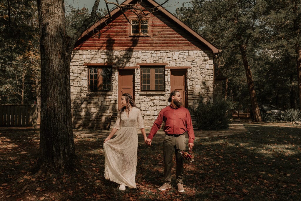 Bride and groom stand in front of a stone building while holding hands. They're looking in opposite directions and the groom is holding the bouquet.