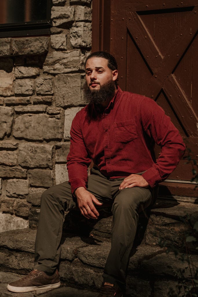 Groom sits on steps outside a stone building. The sun is shining on him.