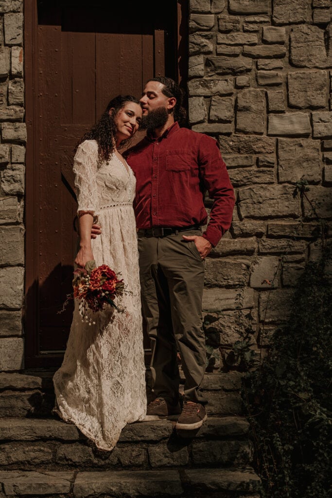 Bride and groom stand on stone steps outside a stone building in front of the door. The groom is kissing her forehead.