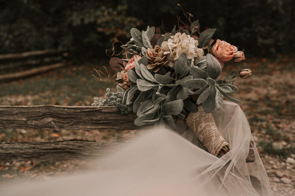 A bridal bouquet rests on top of a veil and a rustic wooden fence.