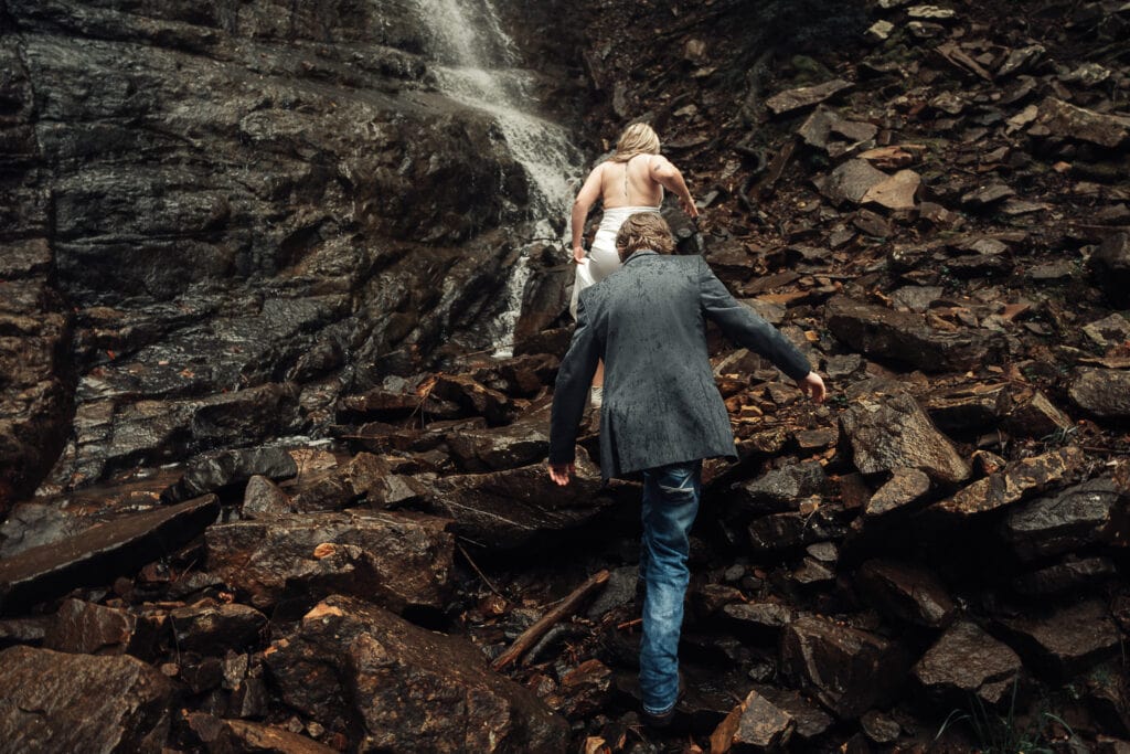 Bride & Groom climb up slippery rocks towards a waterfall. It is raining and couple's wedding attire is wet.