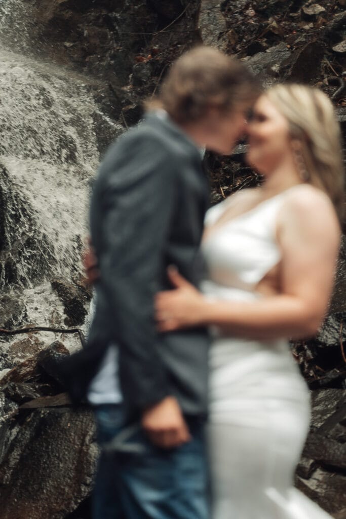 Groom is passionately kissing bride by holding onto sides of her face. They're standing on rocks in front of waterfall.