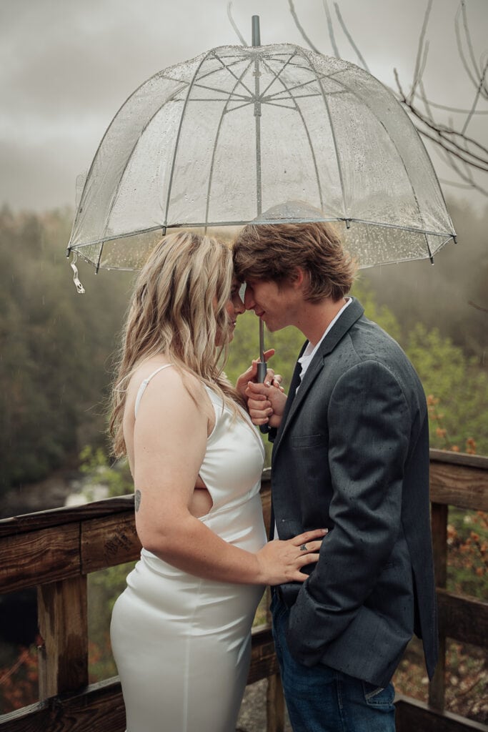 A couple stands on waterfall overlook, nose to nose, under an umbrella.