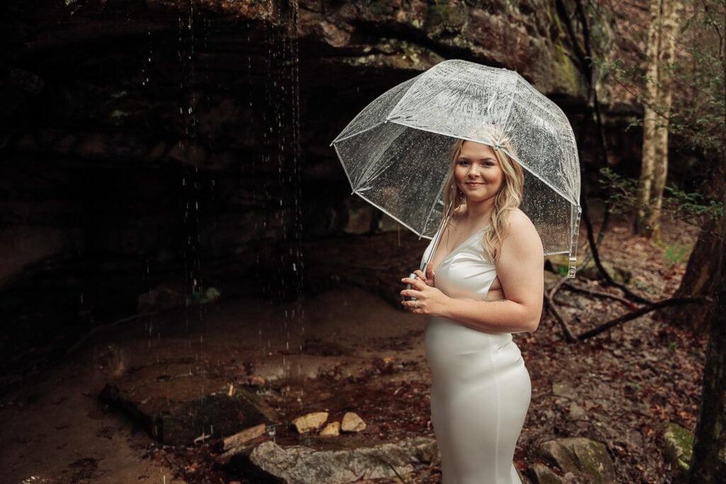 Bride stands off to the side of Fiery Gizzard trail while holding umbrella. Rain water is dripping off rocks behind her
