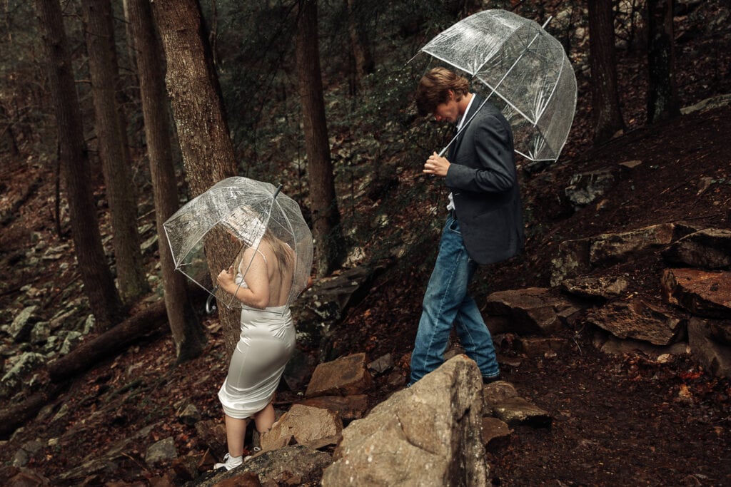 A couple hikes down a muddy trail while holding umbrellas. They're dressed in wedding attire.