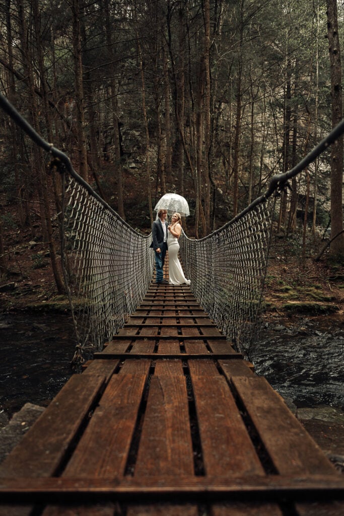 Bride and Groom stand under a clear umbrella on a swinging bridge, during their hiking elopement.