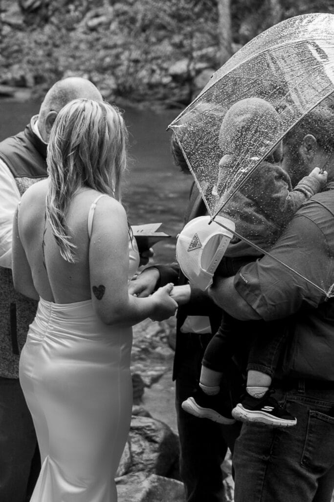 A family holds hands and prays together during an outdoor wedding ceremony in the rain. One person is holding a clear umbrella over the child he's holding.