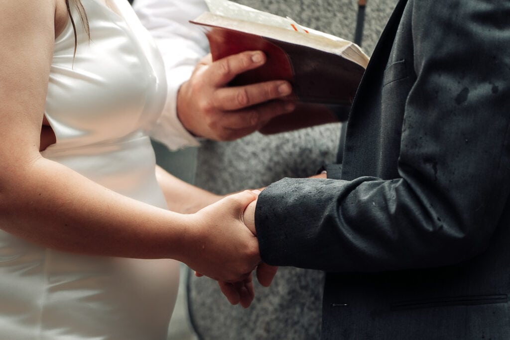 A couple holds hands during their elopement ceremony. The Bible is being held in the background by the officiant.
