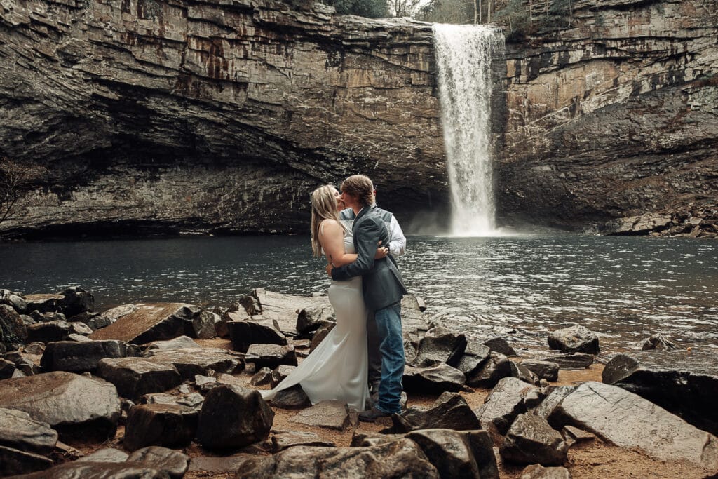 Wide angle image of couple's first kiss. They are standing on rocks. Foster Falls is in the background.