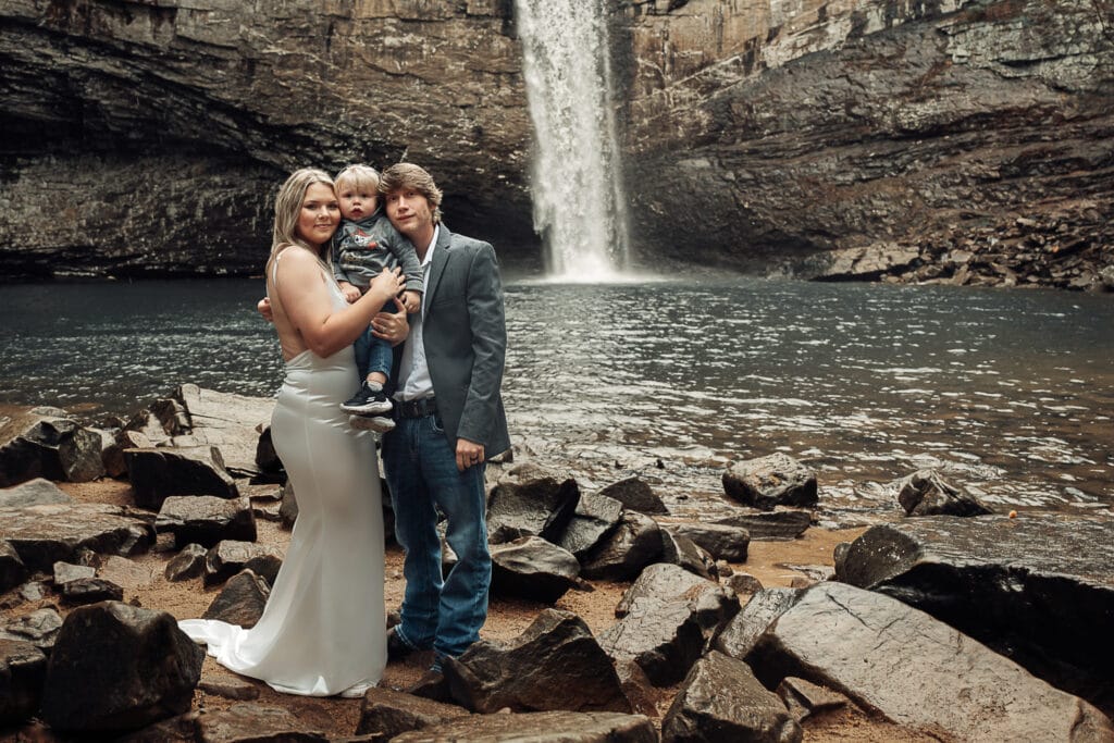 Ashlyn & Brent stand, dressed in wedding attire, in front of the waterfall with their son.