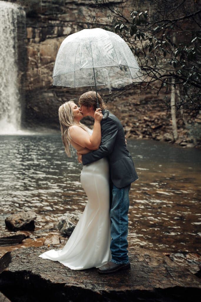 Ashly & Brent hold an umbrella over them as they kiss in front of waterfall in the pouring rain during their elopement