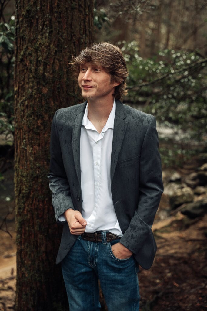 Brent poses for a portrait in his suitcoat and jeans. He's standing next to a tree of to the side of Foster Falls trail.