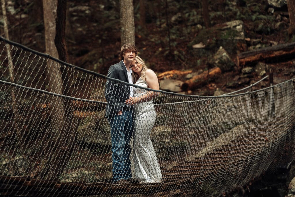 Ashlyn & Brent pause on the middle of the swinging bridge. They're centered in the image with the forest behind them.