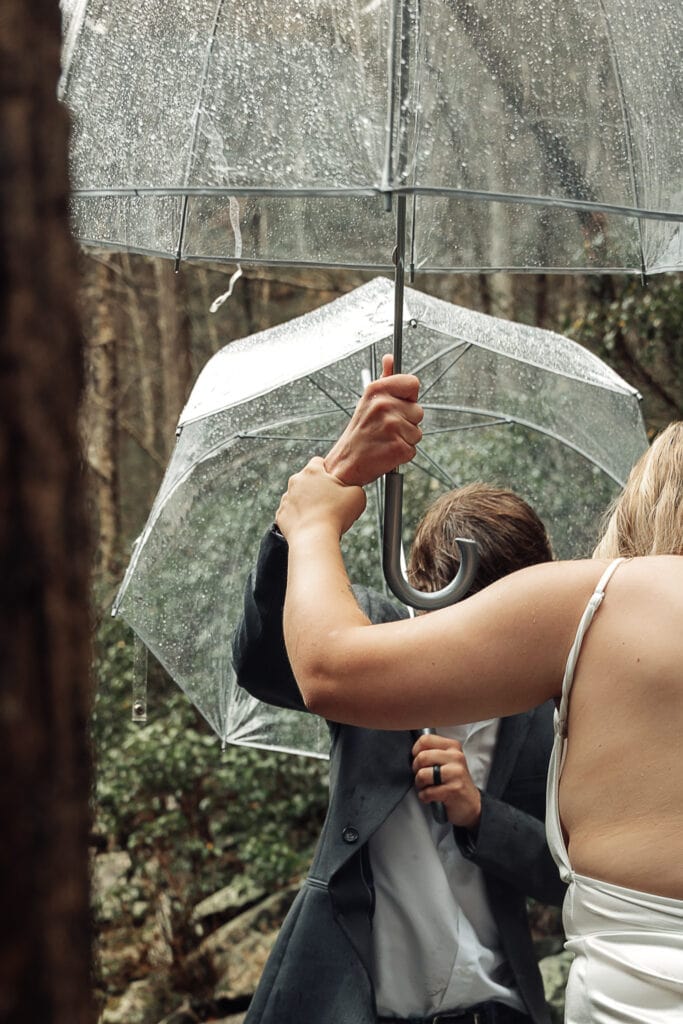 Closeup of Bridge holding onto Groom's arm as they hold umbrellas and he helps her on the muddy trail at Foster Falls.