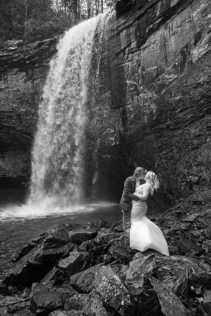 Ashlyn has her arm around Brent as she kisses him while standing on slippery rocks. A waterfall is behind them