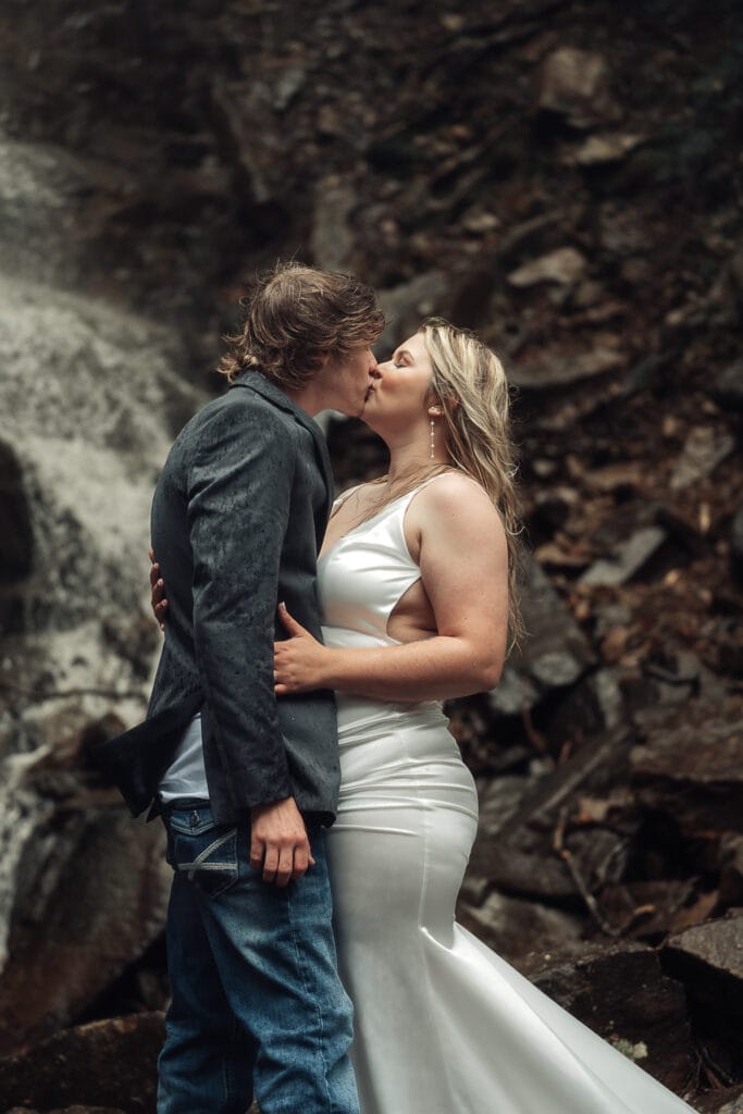 Closeup image of couple in wedding attire kissing at the base of waterfall during the rain.