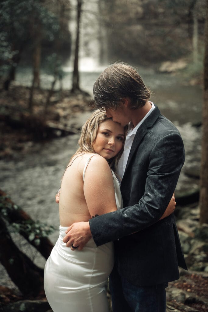 Ashlyn & Brent hug in the rain during their elopement. Brent kisses her forehead. Waterfall is behind them.