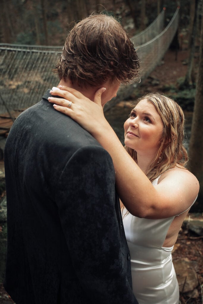 Ashlyn looks up at Brent during their outdoor elopement in the rain. A swinging bridge is in the background.