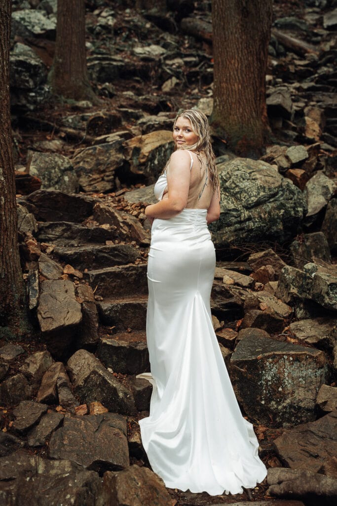Bride pauses in the rain while hiking up rock steps on trail. She looks over her shoulder at the camera.