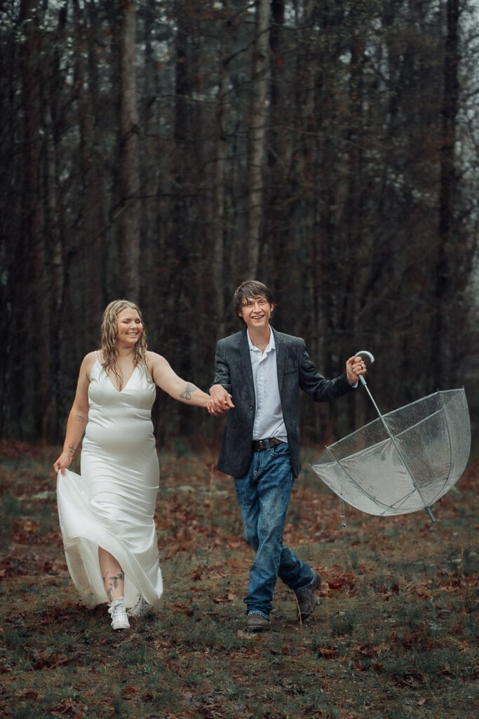 Ashlyn & Brent laugh as they play in the rain on the trail during their elopement. Brent holds umbrella to the side.