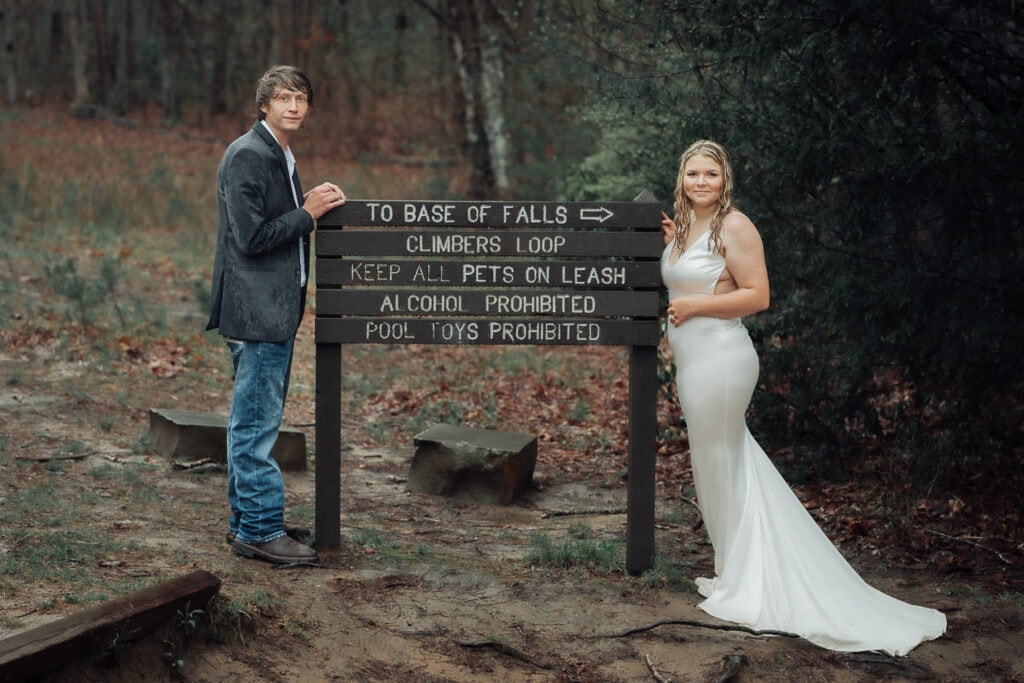 A couple stands on each side of the Foster Falls sign. It's pouring rain and they're in wedding attire.
