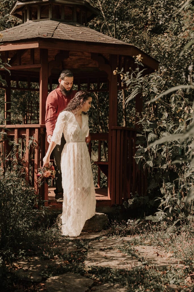 A bride is stepping out of a gazebo to walk down a path while holding hands with the groom who is behind her.