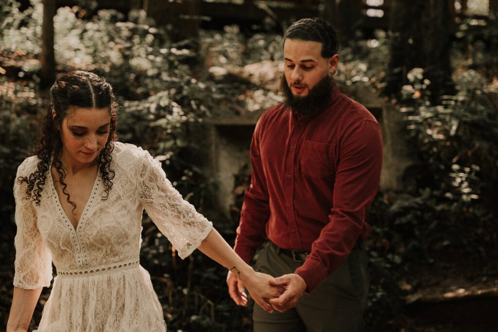 A couple in wedding attire walk hand in hand through the woods.