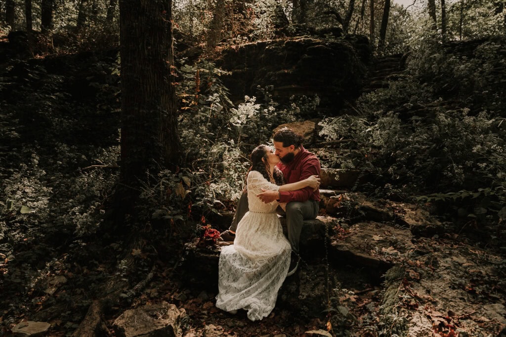 A couple sits on a rock in the forest and kisses as the sun shines on them during their elopement.