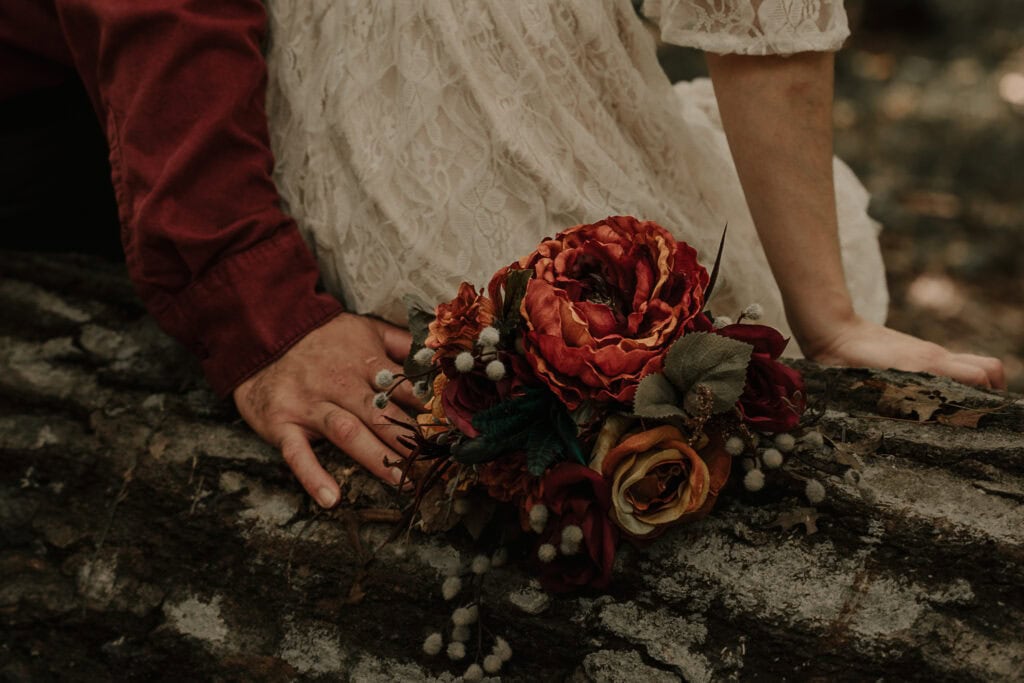 An eloping couple sits on a log. His arm rests behind the bride, resting his hand on her bouquet.