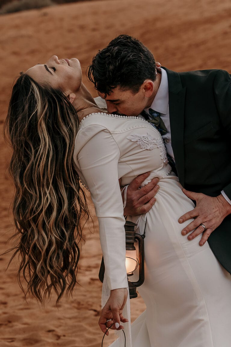 Groom dips the bride as he kisses her chest. She is holding lanterns. They are on sand dunes in Utah.