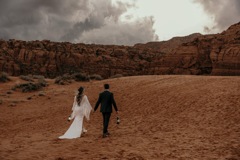 Boho bride and groom walk toward red rock cliffs on sand dunes in Snow Canyon State Park.