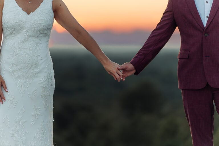 Close up of bride and grooms arms as they hold hands in front of a sunset.