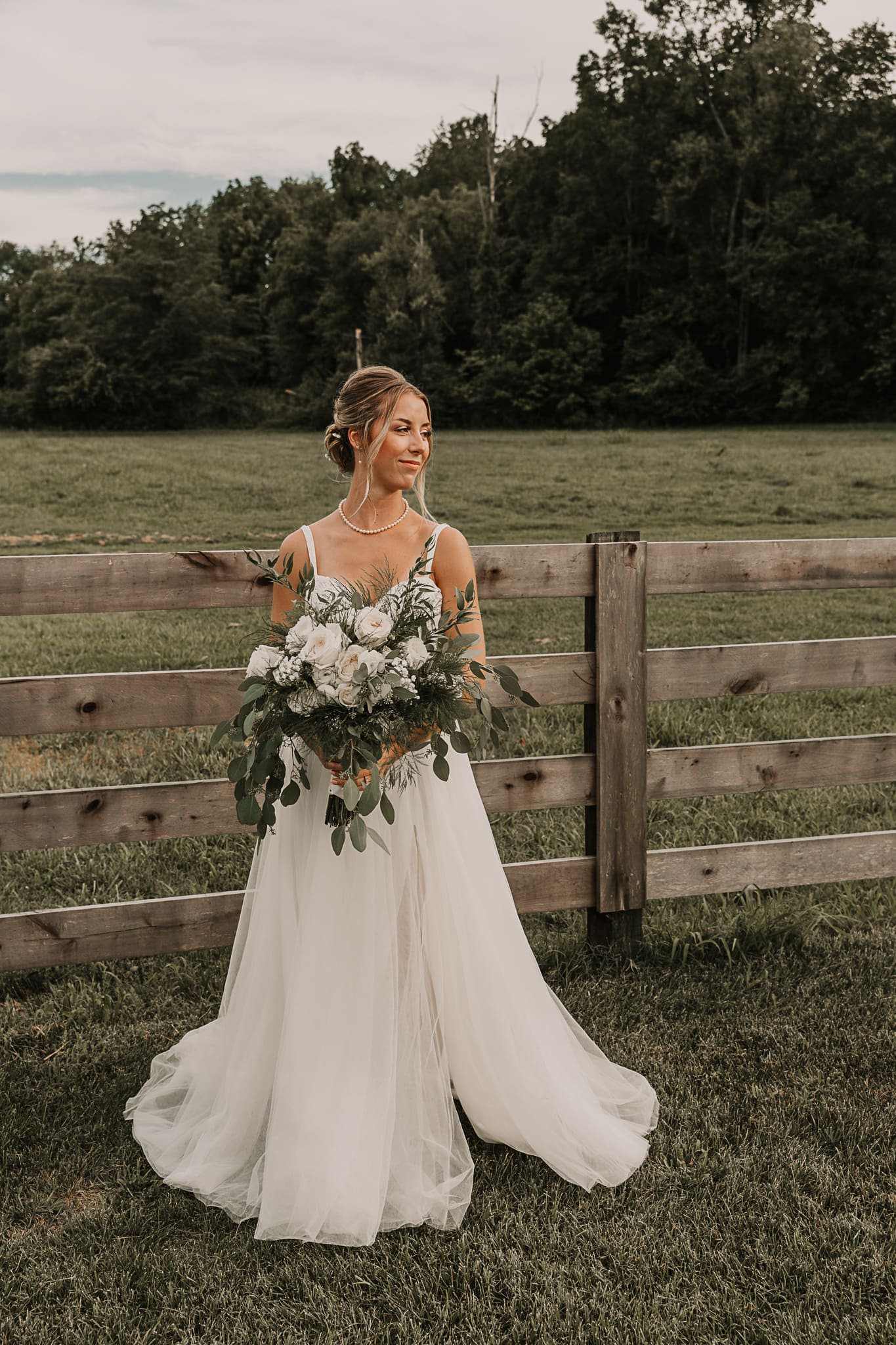 Bride holds bouquet and poses in front of a fence, while looking over her shoulder.