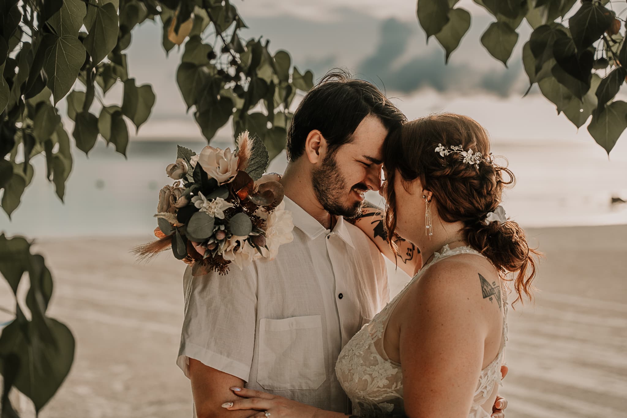Couple stands under a tree branch with their foreheads touching. The ocean is behind them, and they are in wedding attire.