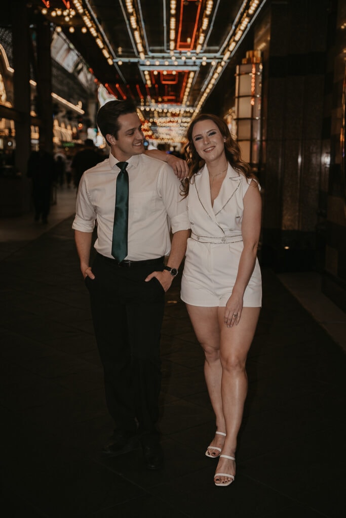 A smiling woman stands with her elbow on mans shoulder while he smiles at her. Vegas lights are in the bacground.