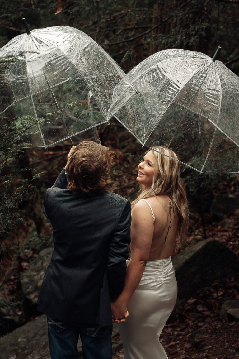 Couple hikes in the rain under umbrellas, dressed in wedding attire, down the Fiery Gizzard Trail.