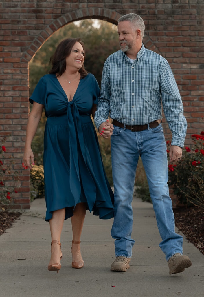 Couple laughs as they walk hand in hand through a botanical garden. A brick archway is in the rose garden behind them.