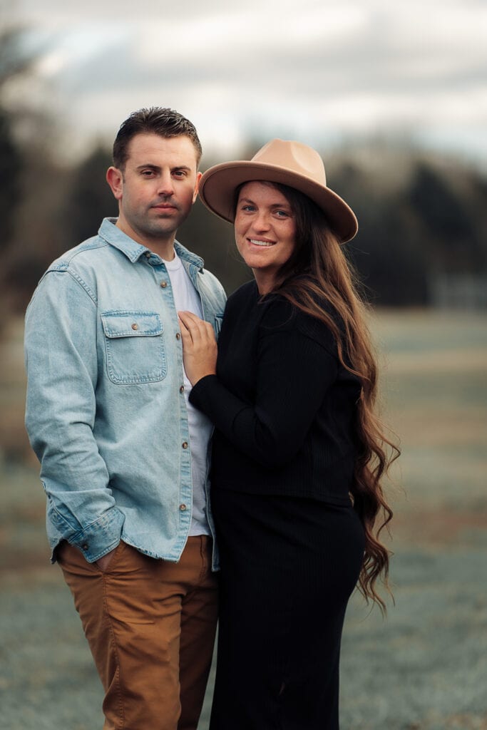 A man and woman stand outdoors and smile at the camera. She is wearing a boho hat and he is dressed in denim.