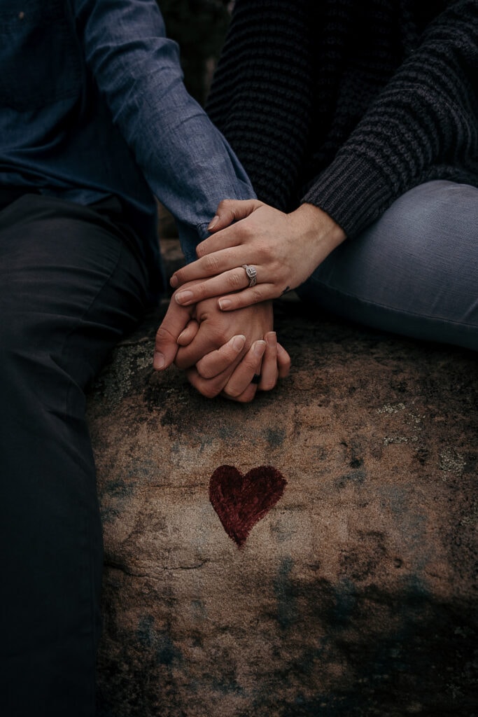 A couple holds hands on a rock outside that has been graffitied with a red heart.