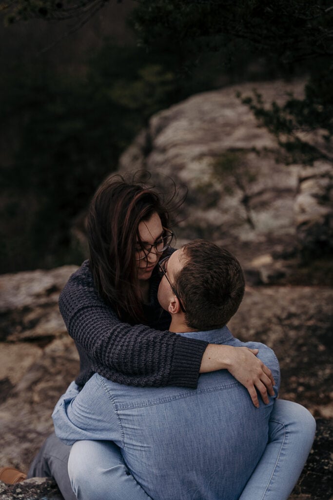 A woman sits on her husband's lap outside on a rock cliff as they look at each other.