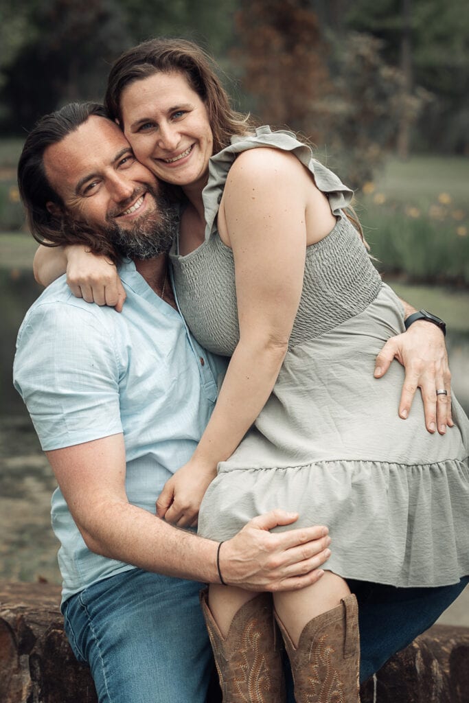 A woman sits on her husbands lap as they laugh while looking at the camera.