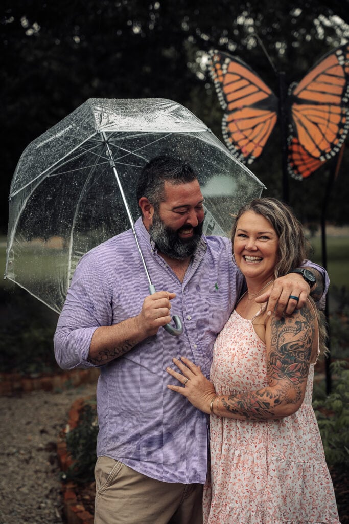 Man holds clear umbrella as he looks at his fiancé during a rainy day photoshoot.